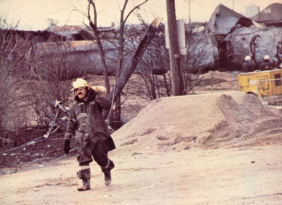 Firefighter carrying extingusher walking with wreckage in the background.